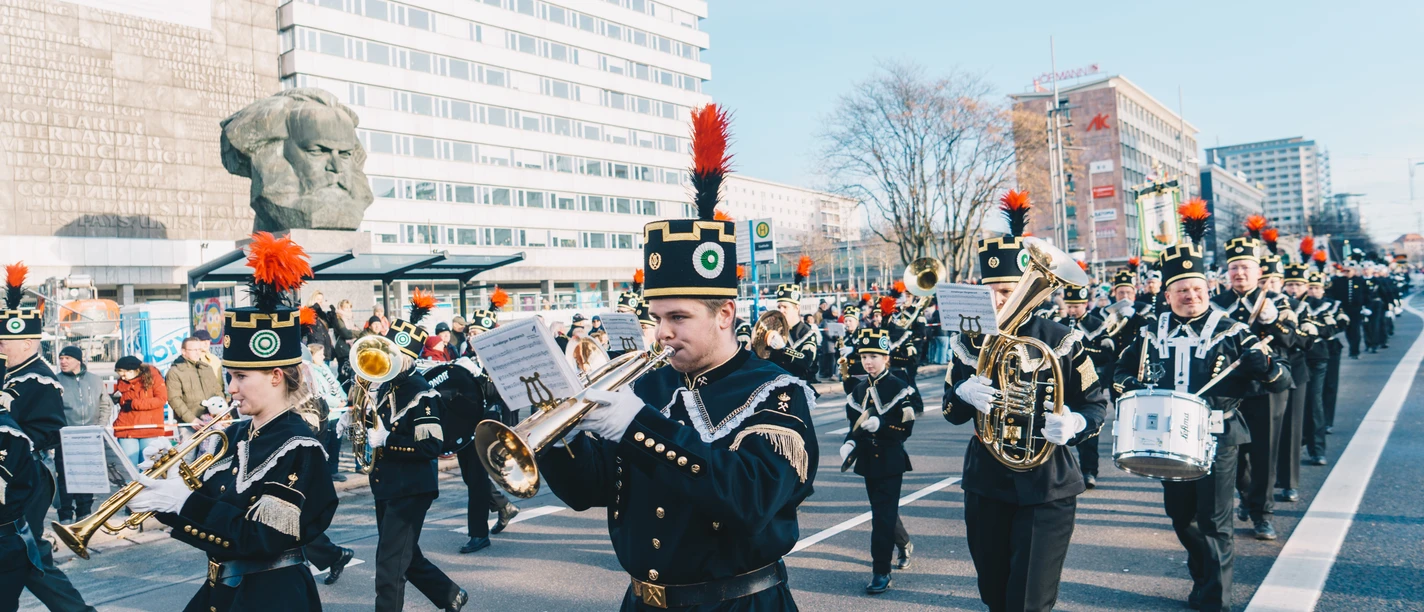 Bergparade vorbei am Karl-Marx-Kopf. © Ernesto Uhlmann_CWE.jpg Musikkapelle in traditioneller Uniform marschiert vor dem Karl-Marx-Denkmal in einer Stadt.