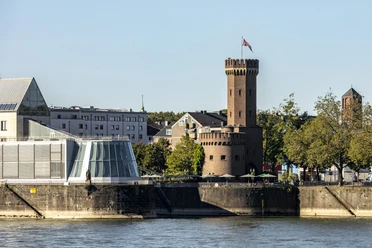 Malakoff Tower The historic Malakoff Tower stands on the banks of the Rhine in Cologne, flanked by modern buildings and surrounded by trees that glow in the afternoon sun.