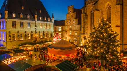 Ein festlich beleuchteter Weihnachtsmarkt bei Dämmerung mit Ständen und einem geschmückten Tannenbaum.