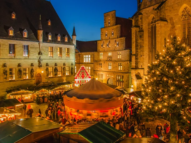 Ein festlich beleuchteter Weihnachtsmarkt bei Dämmerung mit Ständen und einem geschmückten Tannenbaum.