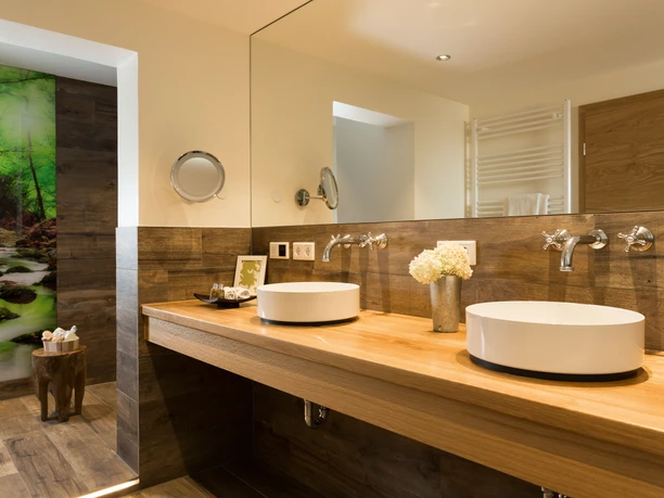 Germanenhof_Sandebeck_JagdSuite_Badezimmer.jpg Luxuriöses Badezimmer mit zwei runden Waschbecken, Holzdetails und Waldmotiv an der Duschwand.
