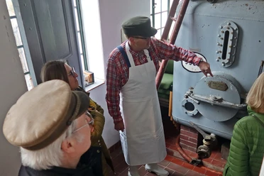 Alte Brennerei szenische Führung mit Erwin.JPG Erwin leitet eine Gruppe durch eine historische Brennerei, zeigt Details eines antiken Kessels.