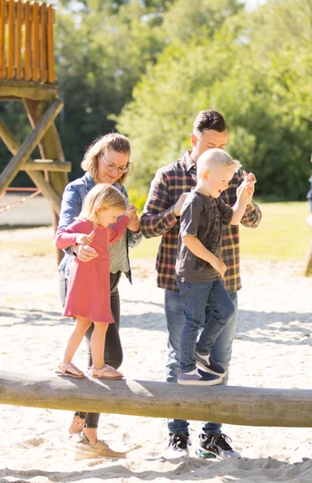 Eine Familie balanciert auf einem Baumstammspielgerät in einem sonnenverwöhnten Spielplatz.