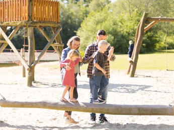 Eine Familie balanciert auf einem Baumstammspielgerät in einem sonnenverwöhnten Spielplatz.