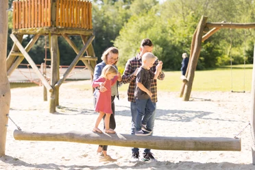 Badesee Mindenerwald Eine Familie balanciert auf einem Baumstammspielgerät in einem sonnenverwöhnten Spielplatz.