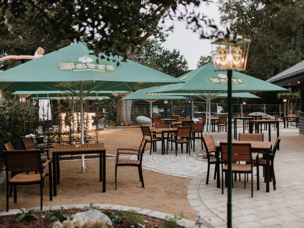 Quiet beer garden with wooden chairs under green parasols in a paved outdoor area.