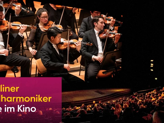 Ein Orchester spielt auf einer Bühne in einem halbdunklen, gut besetzten Konzertsaal.