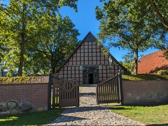 Haus Büter Fachwerkhaus mit steilem Giebel und geöffneter Holztorzufahrt, umgeben von Bäumen und Backsteinmauer.