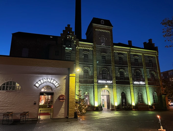 BRAUWELT KÖLN Abendlicher Blick auf eine beleuchtete, historische Backsteinbrauerei mit Eingang zur Brennerei.Evening view of an illuminated, historic brick brewery with entrance to the distillery.
