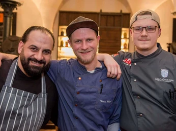 Drei Köche in Arbeitskleidung stehen lächelnd Arm in Arm in einem stilvoll beleuchteten Raum.