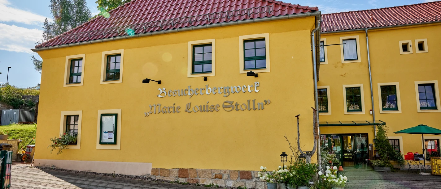 Außenansicht Touristinformation Das Erlebnisbergwerk "Marie Louise Stolln" präsentiert sich mit gelber Fassade und roten Dachziegeln.