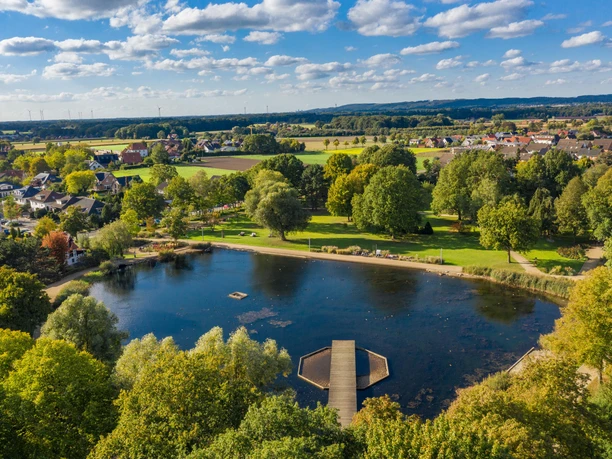Der Kurpark von Bad Laer aus der Vogelperspektive Landschaft mit See, Steg, grünen Bäumen und Dörfern im Hintergrund unter blauem Himmel.