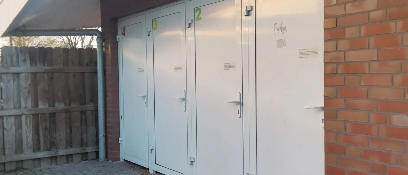 IMG20231128150120.jpg Auf dem Bild sind vier weiße, farbig nummerierte Türen an einem Backsteingebäude zu sehen, auf einem gepflasterten Boden.