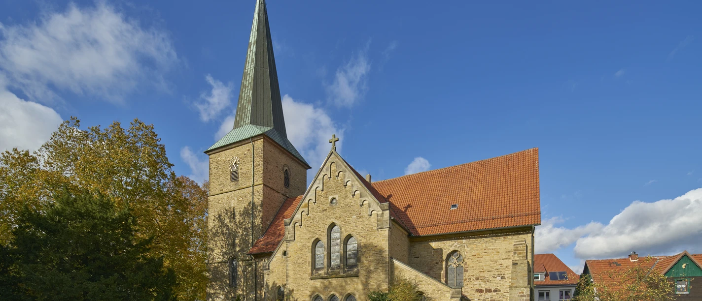 Historische kerk met een spitse toren en rood dak, omringd door bomen onder een blauwe hemel.