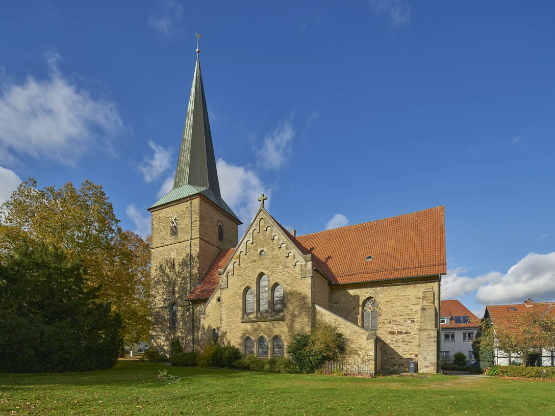 Sankt Laurentius in Bissendorf Historische Kirche mit spitzem Turm und rotem Dach, umgeben von Bäumen unter blauem Himmel.Historic church with a pointed tower and red roof, surrounded by trees under a blue sky.Historisk kirke med spidst tårn og rødt tag, omgivet af træer under en blå himmel.Historische kerk met een spitse toren en rood dak, omringd door bomen onder een blauwe hemel.
