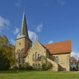 Sankt Laurentius in Bissendorf Historische Kirche mit spitzem Turm und rotem Dach, umgeben von Bäumen unter blauem Himmel.Historic church with a pointed tower and red roof, surrounded by trees under a blue sky.Historisk kirke med spidst tårn og rødt tag, omgivet af træer under en blå himmel.Historische kerk met een spitse toren en rood dak, omringd door bomen onder een blauwe hemel.