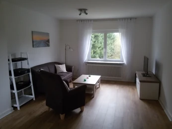 Ferienwohnung Niebaum Wohnzimmer Helles Wohnzimmer mit großem Fenster, braunem Sofa, Sessel, Couchtisch und TV, mit Blick ins Grüne.