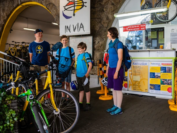 Cologne bike station Four people in casual clothes, with bicycles in front of an information desk.