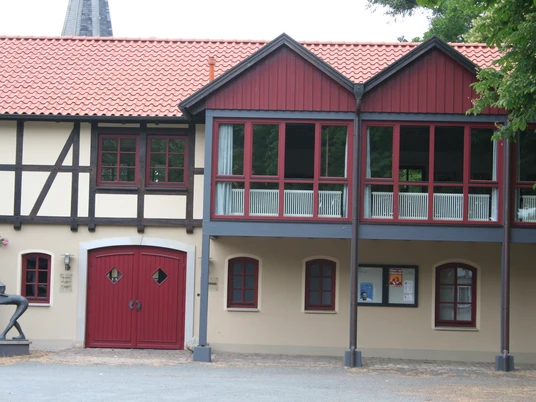 Bürgerhaus Uchte Fachwerkgebäude mit rotem Ziegeldach und Holzverzierungen, darüber befindet sich ein verglaster Balkon.