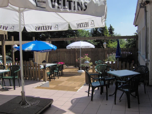 Biergarten Stille Ein Biergarten mit Holzzaun, grünen Stühlen und Sonnenschirmen, umgeben von Bäumen und Blumen.