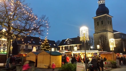 Abendlicher Weihnachtsmarkt mit Buden, geschmücktem Baum und im Hintergrund eine Kirche mit Turm.