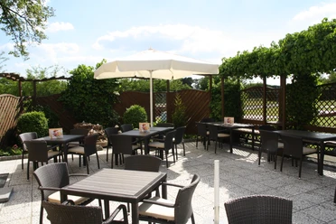 Sonnige Terrasse eines Cafés mit dunklen Tischen und Stühlen, umgeben von grüner Pflanzenwand.