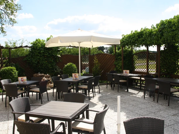 Sonnige Terrasse eines Cafés mit dunklen Tischen und Stühlen, umgeben von grüner Pflanzenwand.
