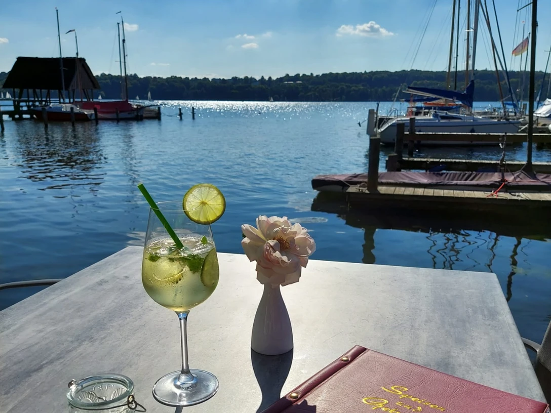 Restaurant.jpg Blick vom Ufer auf den See, im Vordergrund ein Tisch mit einem Cocktail und einer Speisekarte darauf.