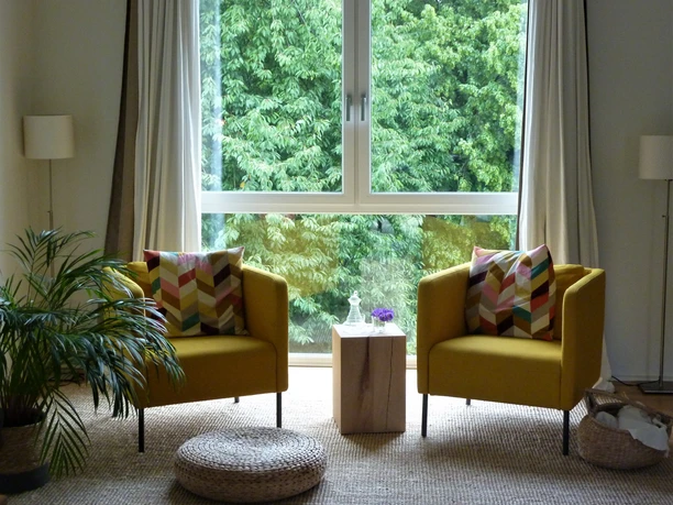 Ferienwohnung Zeitraum Zwei gelbe Sessel mit bunten Kissen stehen vor einem großen Fenster mit Blick auf grüne Bäume.