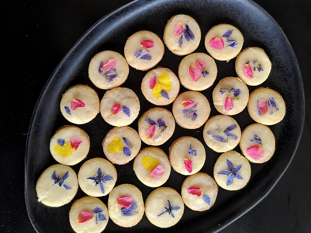 Bluetenkekse Ein schwarzer Teller mit Keksen, verziert mit essbaren Blüten, auf dunklem Hintergrund.