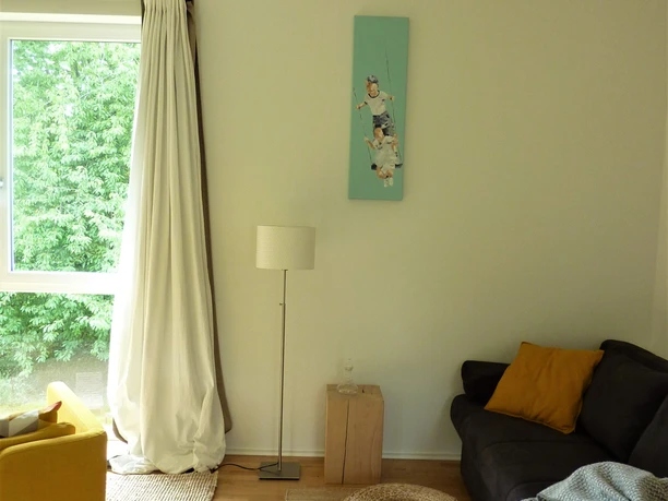 Ferienwohnung Zeitraum Helles Wohnzimmer mit gelbem Sessel, grauem Sofa, hellen Vorhängen und dekorativem Teppich.