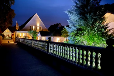 Wilsmann Apartments Hövelhof bei Nacht, beleuchtet mit umgebenden Pflanzen und einem dekorativen Zaun.