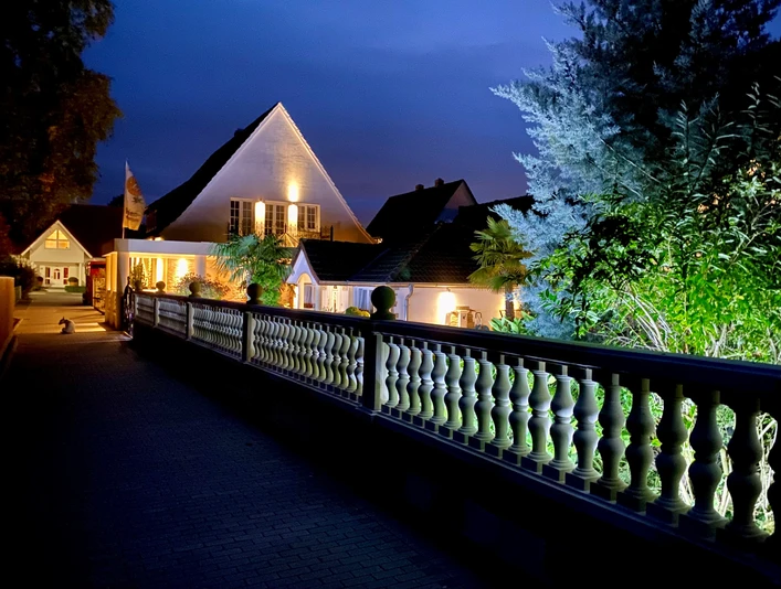 Wilsmann Apartments Hövelhof bei Nacht, beleuchtet mit umgebenden Pflanzen und einem dekorativen Zaun.