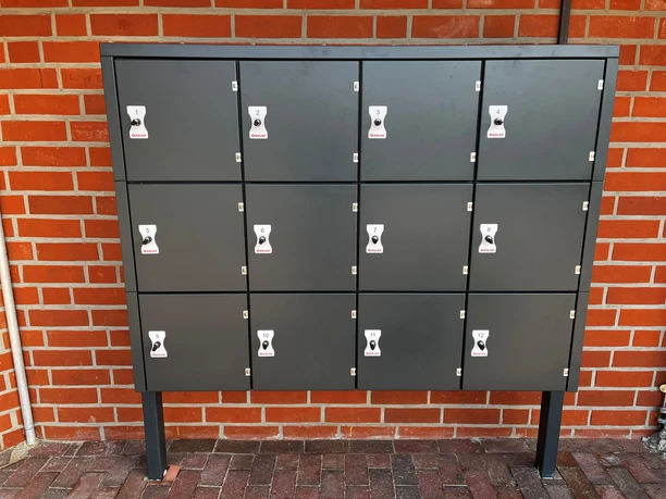 Fahrradschließanlage Greetsiel Grau-schwarze Fahrradschließfächer mit zwölf nummerierten Türen vor roter Ziegelwand in Greetsiel.