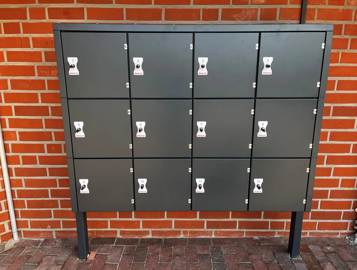 Fahrradschließanlage Greetsiel Grau-schwarze Fahrradschließfächer mit zwölf nummerierten Türen vor roter Ziegelwand in Greetsiel.