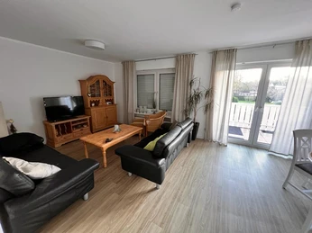 FerienwohnungNeuwöhner_WohnzimmerCC-BY-SA_Teutoburger_Wald_Stadt_Schloss_Holte-Stukenbrock (9).jpg Modernes Wohnzimmer mit schwarzen Ledersofas, Holzmöbeln, großem Fenster und Zugang zum Balkon.