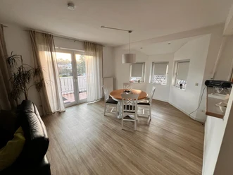 Esszimmer mit rundem Holztisch, vier weißen Stühlen, großer Fensterfront und Zugang zu Balkon.