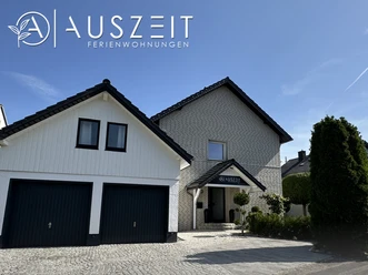 Modernes zweistöckiges Haus mit zwei Garagen, umgeben von gepflegter Landschaft und blauem Himmel.