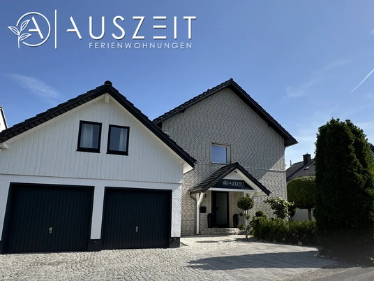 FerienwohnungA'USZEIT_AußenansichtCC-BY-SA_Teutoburger_Wald_Stadt_Schloss_Holte-Stukenbrock.jpg Modernes zweistöckiges Haus mit zwei Garagen, umgeben von gepflegter Landschaft und blauem Himmel.