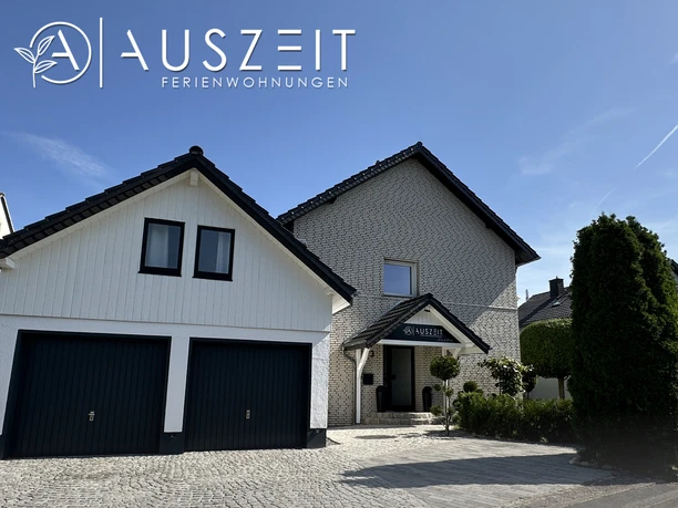 FerienwohnungA'USZEIT_AußenansichtCC-BY-SA_Teutoburger_Wald_Stadt_Schloss_Holte-Stukenbrock.jpg Modernes zweistöckiges Haus mit zwei Garagen, umgeben von gepflegter Landschaft und blauem Himmel.