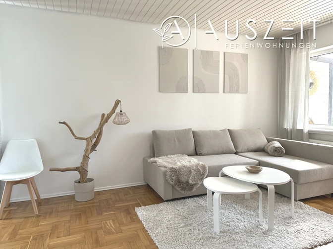 Helles Wohnzimmer einer Ferienwohnung mit grauem Sofa, weißen Tischen und großen Fenstern.
