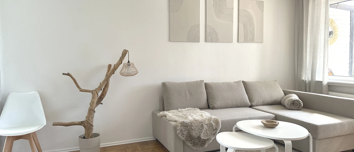 Helles Wohnzimmer einer Ferienwohnung mit grauem Sofa, weißen Tischen und großen Fenstern.