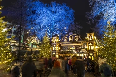 Heinzels Wintermärchen Der Weihnachtsmarkt auf dem Alter Markt in Köln strahlt im Lichterglanz mit Festdekoration und festlichem Torbogen, eingerahmt von blauen Bäumen.
