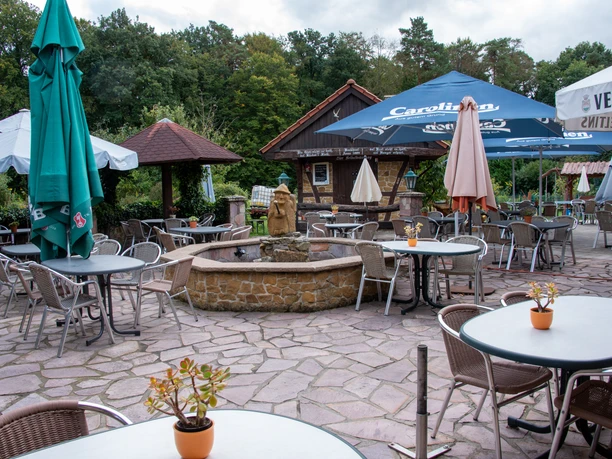 Kaffee Hexenbrink Biergarten Ein idyllisches Außencafé mit Steinbrunnen, Tischen und Schirmen auf gepflastertem Untergrund.