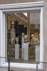 Belgisches Haus RGM In einem großen Fenster eines historischen Gebäudes sind mehrere antike Büsten und Skulpturen auf eleganten Sockeln ausgestellt. Die steinernen Kunstwerke variieren in Form und Größe, strahlen zeitlose Eleganz aus und werden durch eine helle Beleuchtung akzentuiert.