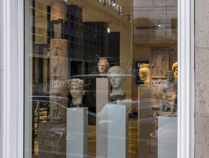 Belgisches Haus RGM In einem großen Fenster eines historischen Gebäudes sind mehrere antike Büsten und Skulpturen auf eleganten Sockeln ausgestellt. Die steinernen Kunstwerke variieren in Form und Größe, strahlen zeitlose Eleganz aus und werden durch eine helle Beleuchtung akzentuiert.In a large window of a historic building, several antique busts and sculptures are displayed on elegant pedestals. The stone works of art vary in shape and size, exude timeless elegance and are accentuated by bright lighting.