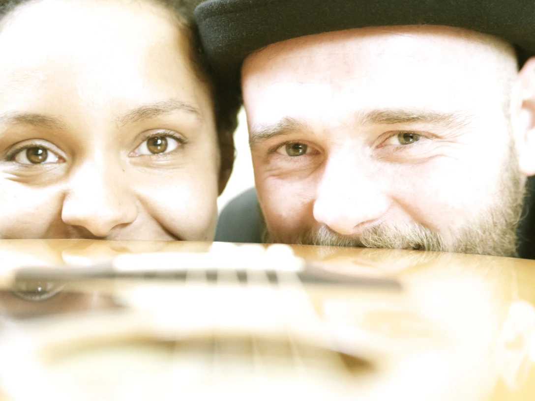 Die Gruppe White Coffee Zwei Menschen lächeln hinter einer akustischen Gitarre hervor, die den Vordergrund des Bildes dominiert.