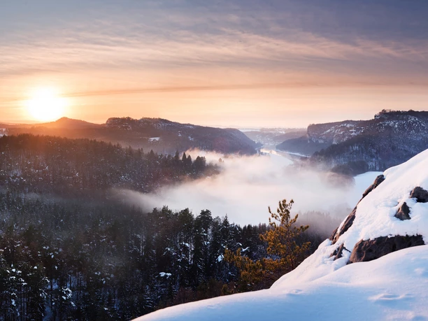 Blick vom Gamrig auf den Kurort Rathen Winterliche Landschaft mit schneebedeckten Bergen und Wäldern im Vordergrund, Nebel über dem Tal und einem Sonnenuntergang am Horizont.