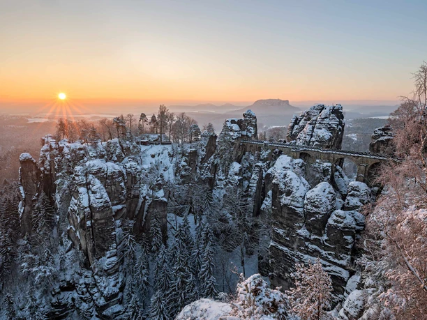 Besneeuwde rotsformaties en een stenen brug bij zonsopgang, omringd door besneeuwde bomen, creëren een vredig winterlandschap.