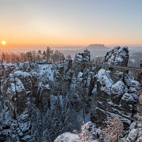 Blick von der Ferdinandaussicht auf die Basteibrücke im Winter Verschneite Felsformationen und eine Steinbrücke im Sonnenaufgang, umgeben von schneebedeckten Bäumen, schaffen eine friedliche Winterlandschaft.